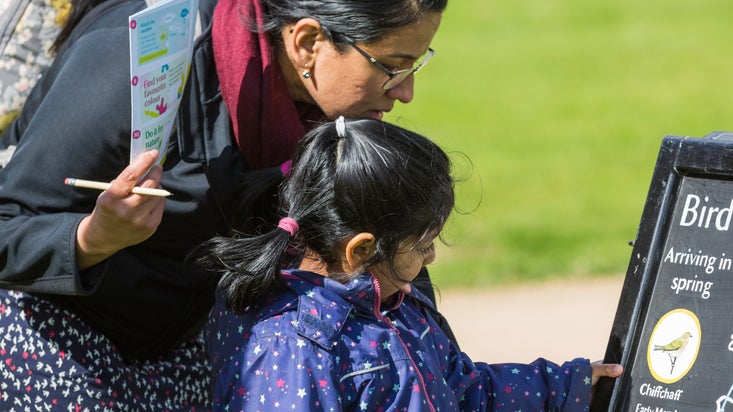 A close up view of a woman and child on an Easter trail looking at a chalkboard in a sunny garden; the woman holds a colourful piece of paper and a pencil